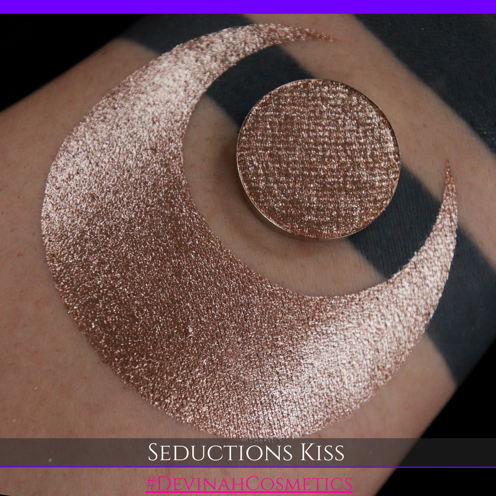 A round eyeshadow pan labeled 'SEDUCTIONS KISS' made by Devinah Cosmetics, placed over a large crescent moon swatch of the same shimmering shifty eyeshadow on skin. The eyeshadow has colors brown, tan, creme sparkle shimmer.