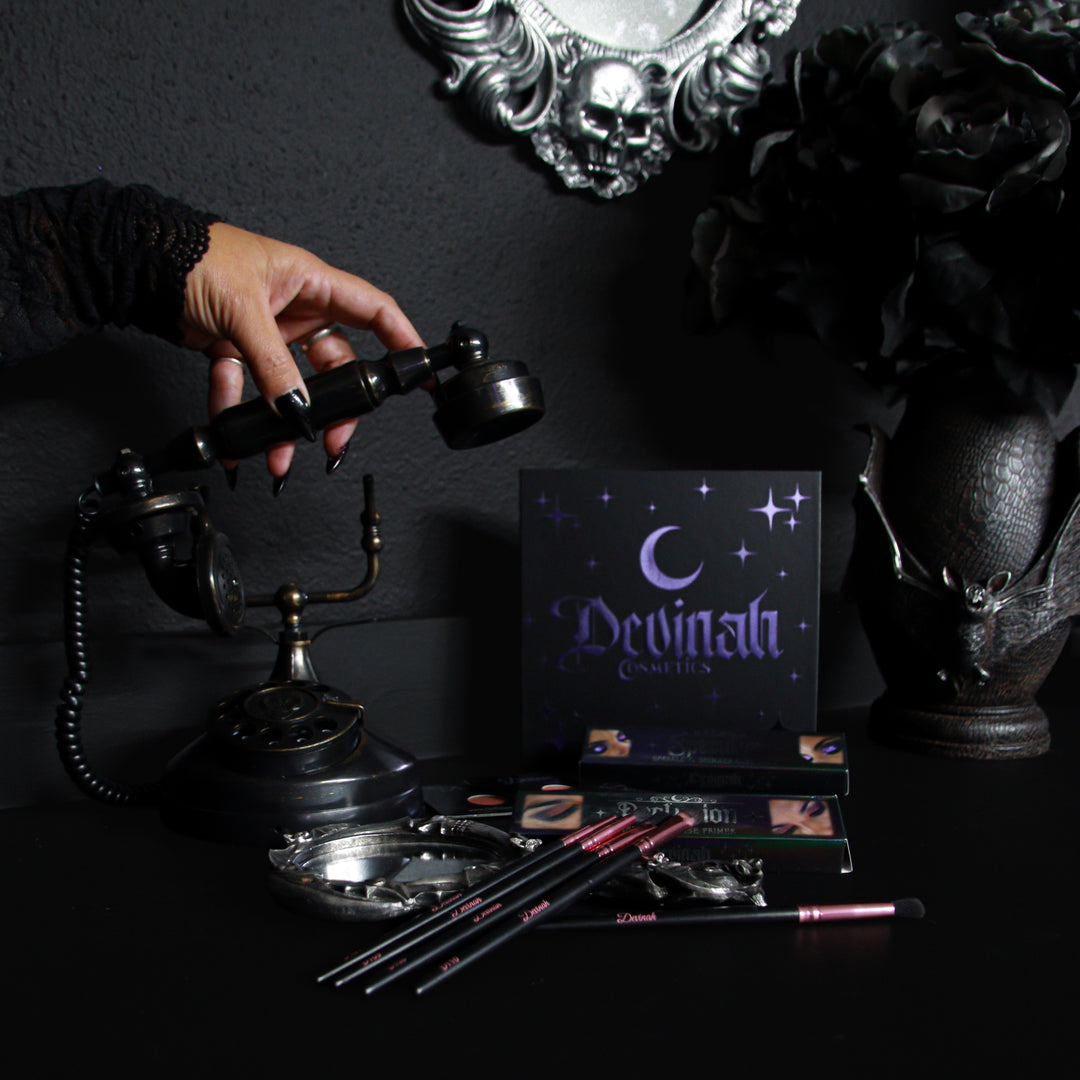 A gothic-themed makeup palette by Devinah Cosmetics displayed on a dark table next to a vintage rotary phone, with a hand holding the phone receiver. The palette packaging is black with purple celestial graphics, and the makeup brushes have pink and silver accents.