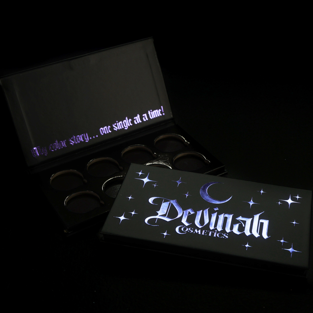 Two black makeup palettes from Devinah Cosmetics on a black background, one open showing circular pans and the other closed with glowing purple gothic lettering and stars with a crescent moon.