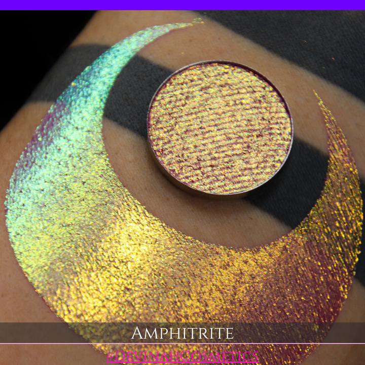 A round eyeshadow pan labeled 'AMPHITRITE' made by Devinah Cosmetics, placed over a large crescent moon swatch of the same shimmering shifty eyeshadow on skin. The eyeshadow has a shift between colors green, gold, purple, blue.