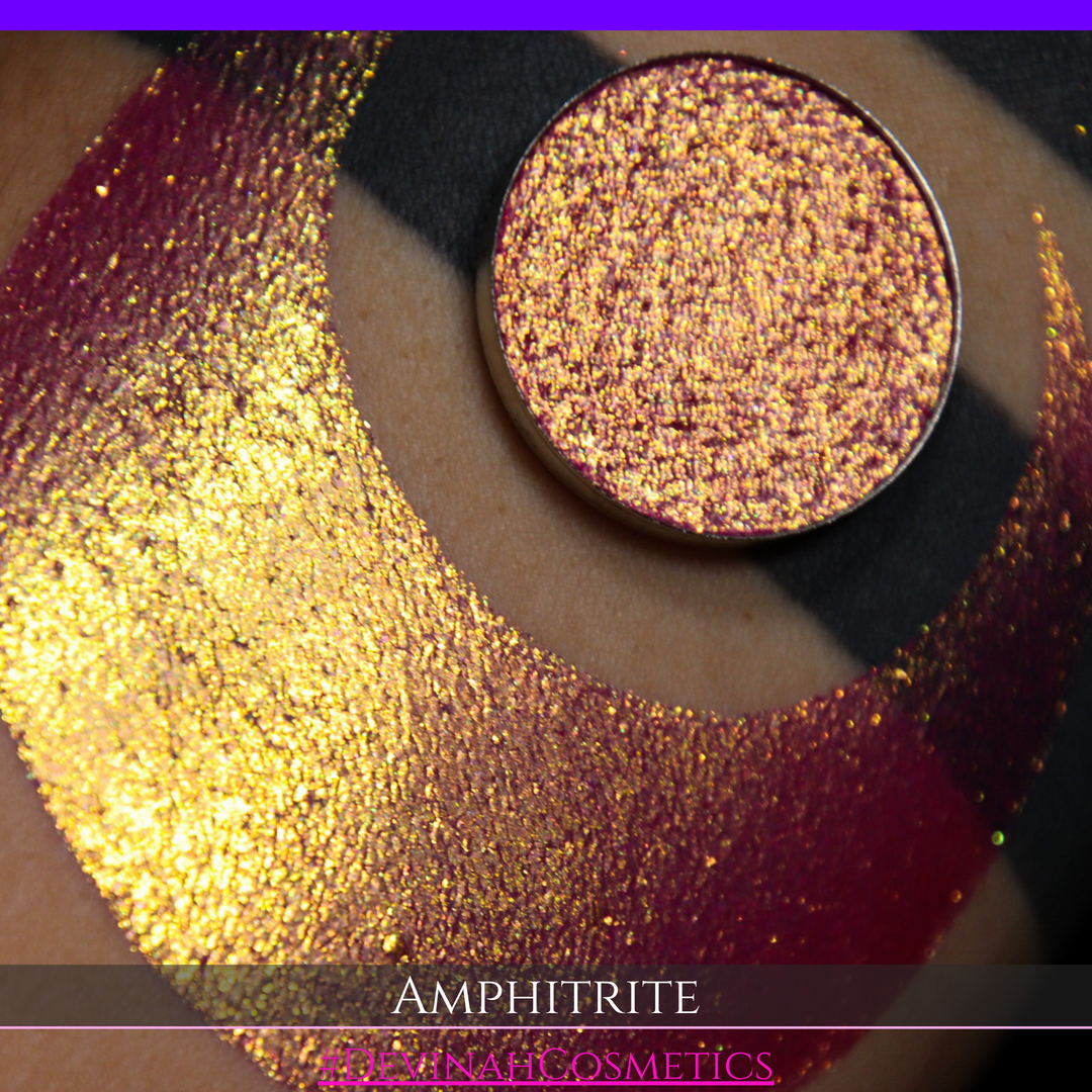 A round eyeshadow pan labeled 'AMPHITRITE' made by Devinah Cosmetics, placed over a large crescent moon swatch of the same shimmering shifty eyeshadow on skin. The eyeshadow has a shift between colors green, gold, purple, blue.