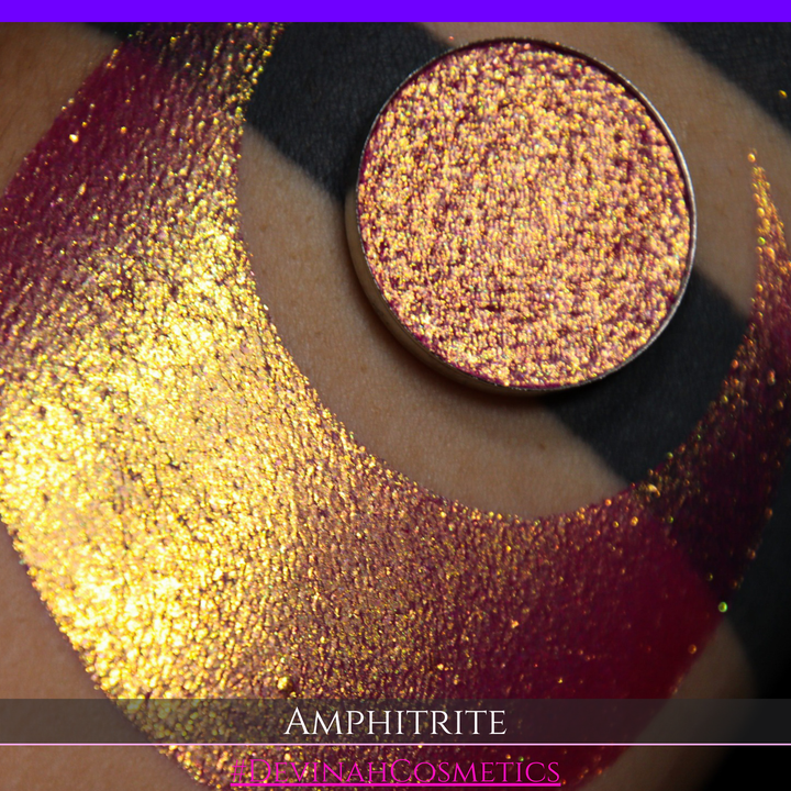 A round eyeshadow pan labeled 'AMPHITRITE' made by Devinah Cosmetics, placed over a large crescent moon swatch of the same shimmering shifty eyeshadow on skin. The eyeshadow has a shift between colors green, gold, purple, blue.