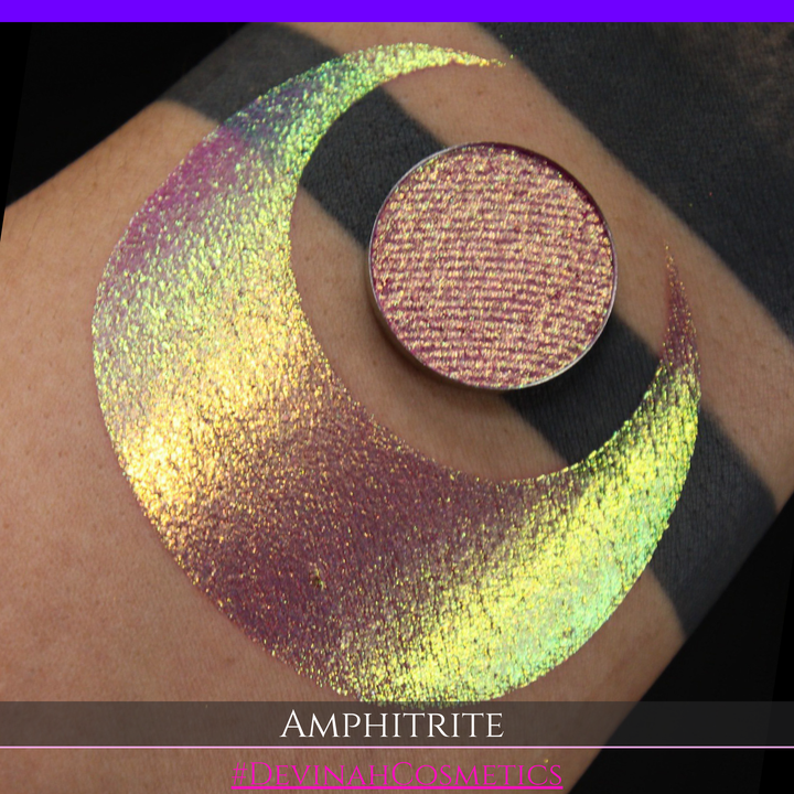 A round eyeshadow pan labeled 'AMPHITRITE' made by Devinah Cosmetics, placed over a large crescent moon swatch of the same shimmering shifty eyeshadow on skin. The eyeshadow has a shift between colors green, gold, purple, blue.