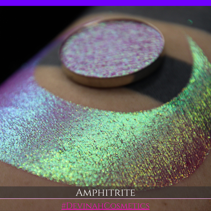 A round eyeshadow pan labeled 'AMPHITRITE' made by Devinah Cosmetics, placed over a large crescent moon swatch of the same shimmering shifty eyeshadow on skin. The eyeshadow has a shift between colors green, gold, purple, blue.
