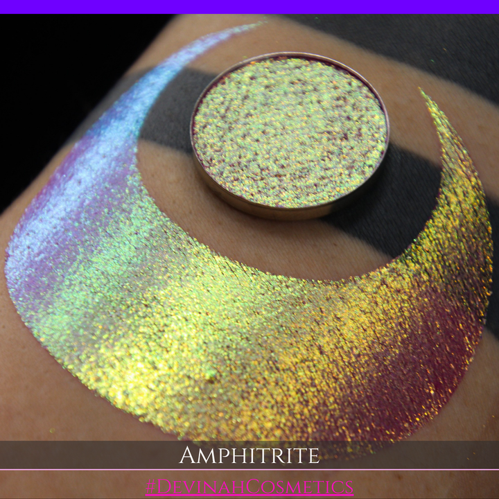 A round eyeshadow pan labeled 'AMPHITRITE' made by Devinah Cosmetics, placed over a large crescent moon swatch of the same shimmering shifty eyeshadow on skin. The eyeshadow has a shift between colors green, gold, purple, blue.