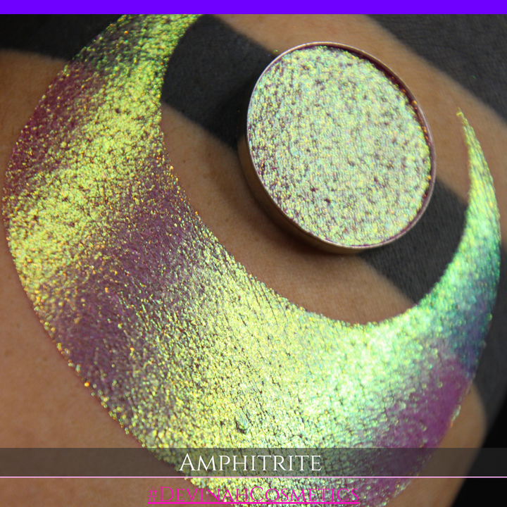 A round eyeshadow pan labeled 'AMPHITRITE' made by Devinah Cosmetics, placed over a large crescent moon swatch of the same shimmering shifty eyeshadow on skin. The eyeshadow has a shift between colors green, gold, purple, blue.