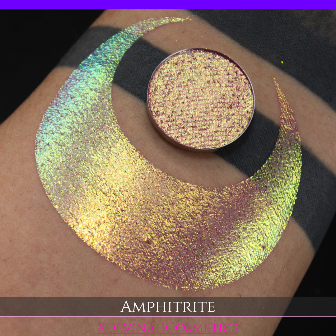 A round eyeshadow pan labeled 'AMPHITRITE' made by Devinah Cosmetics, placed over a large crescent moon swatch of the same shimmering shifty eyeshadow on skin. The eyeshadow has a shift between colors green, gold, purple, blue.