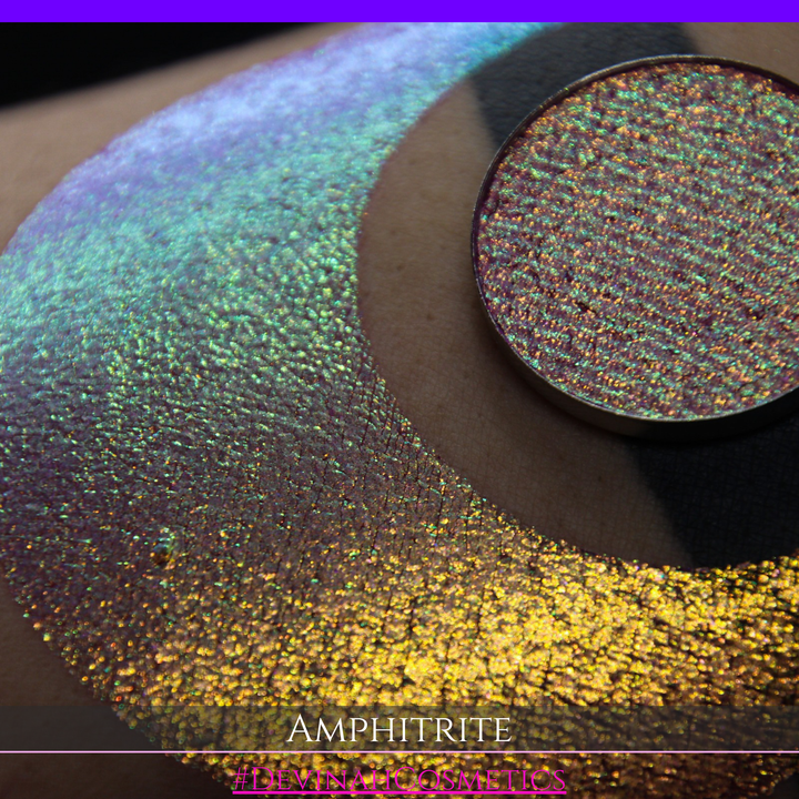 A round eyeshadow pan labeled 'AMPHITRITE' made by Devinah Cosmetics, placed over a large crescent moon swatch of the same shimmering shifty eyeshadow on skin. The eyeshadow has a shift between colors green, gold, purple, blue.