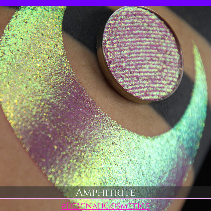 A round eyeshadow pan labeled 'AMPHITRITE' made by Devinah Cosmetics, placed over a large crescent moon swatch of the same shimmering shifty eyeshadow on skin. The eyeshadow has a shift between colors green, gold, purple, blue.