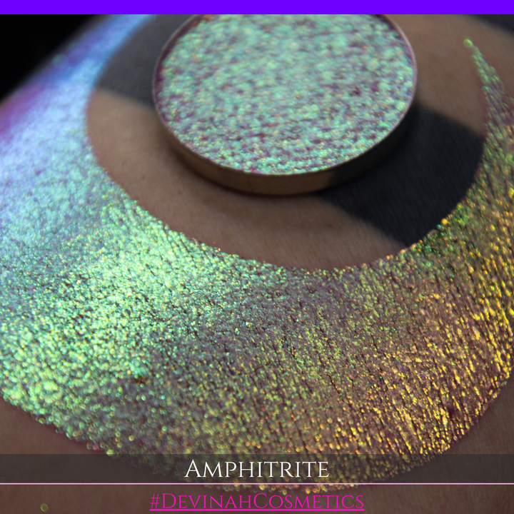 A round eyeshadow pan labeled 'AMPHITRITE' made by Devinah Cosmetics, placed over a large crescent moon swatch of the same shimmering shifty eyeshadow on skin. The eyeshadow has a shift between colors green, gold, purple, blue.