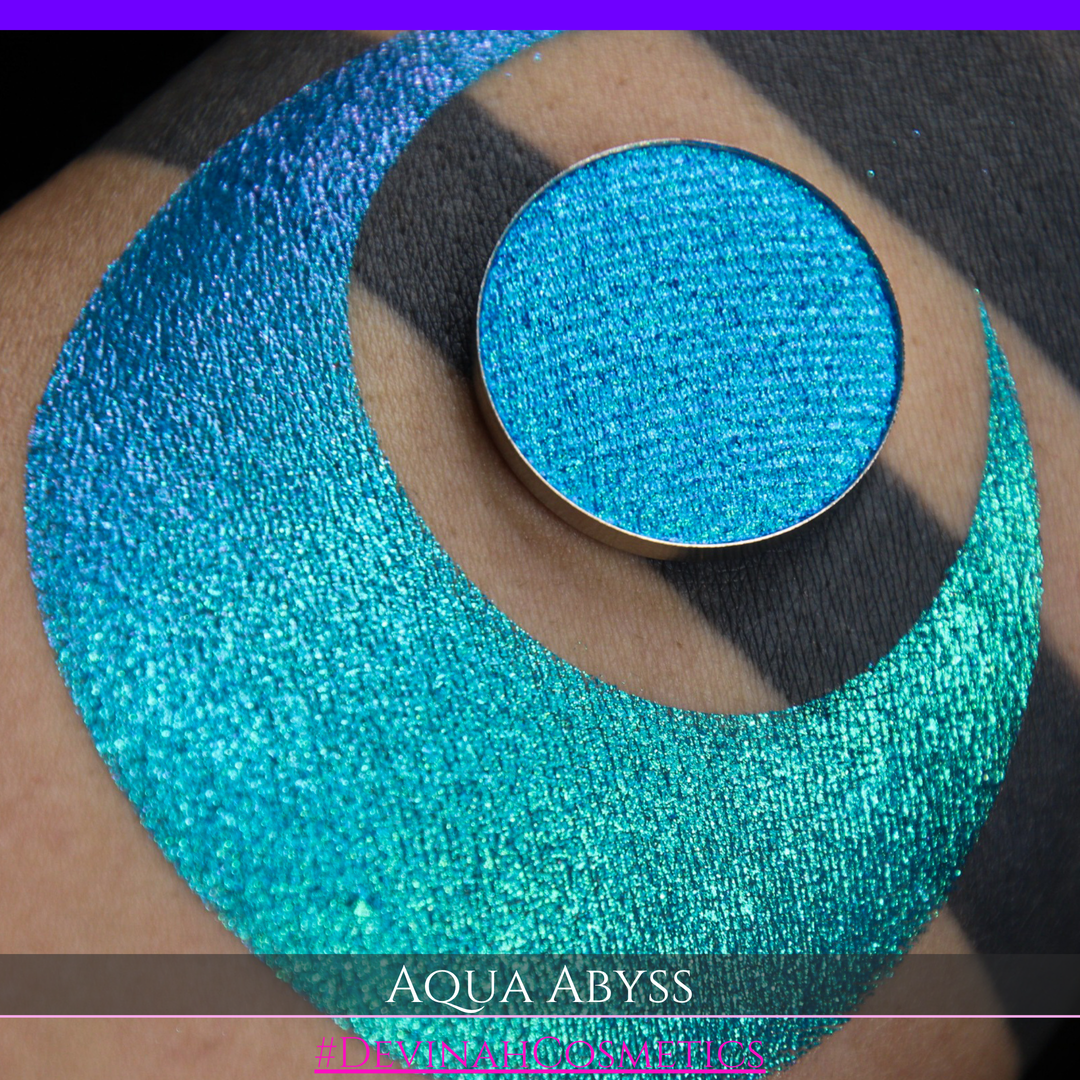 A round eyeshadow pan labeled 'AQUA ABYSS' made by Devinah Cosmetics, placed over a large crescent moon swatch of the same shimmering shifty eyeshadow on skin. The eyeshadow has a duochrome multichrome shift between colors lime green, blue, teal, pink.