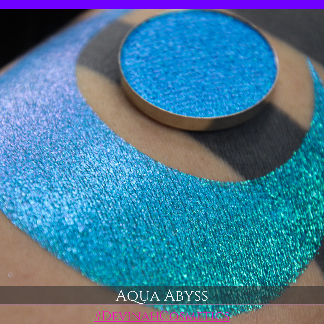 A round eyeshadow pan labeled 'AQUA ABYSS' made by Devinah Cosmetics, placed over a large crescent moon swatch of the same shimmering shifty eyeshadow on skin. The eyeshadow has a duochrome multichrome shift between colors lime green, blue, teal, pink.