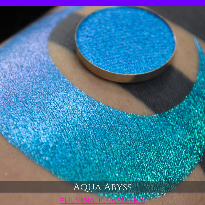 A round eyeshadow pan labeled 'AQUA ABYSS' made by Devinah Cosmetics, placed over a large crescent moon swatch of the same shimmering shifty eyeshadow on skin. The eyeshadow has a duochrome multichrome shift between colors lime green, blue, teal, pink.