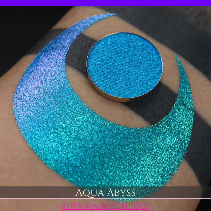 A round eyeshadow pan labeled 'AQUA ABYSS' made by Devinah Cosmetics, placed over a large crescent moon swatch of the same shimmering shifty eyeshadow on skin. The eyeshadow has a duochrome multichrome shift between colors lime green, blue, teal, pink.