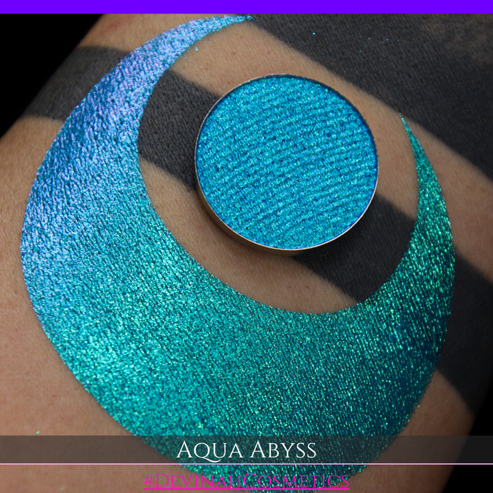 A round eyeshadow pan labeled 'AQUA ABYSS' made by Devinah Cosmetics, placed over a large crescent moon swatch of the same shimmering shifty eyeshadow on skin. The eyeshadow has a duochrome multichrome shift between colors lime green, blue, teal, pink.