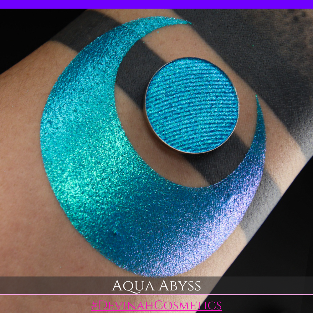 A round eyeshadow pan labeled 'AQUA ABYSS' made by Devinah Cosmetics, placed over a large crescent moon swatch of the same shimmering shifty eyeshadow on skin. The eyeshadow has a duochrome multichrome shift between colors lime green, blue, teal, pink.