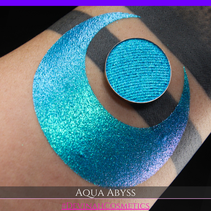 A round eyeshadow pan labeled 'AQUA ABYSS' made by Devinah Cosmetics, placed over a large crescent moon swatch of the same shimmering shifty eyeshadow on skin. The eyeshadow has a duochrome multichrome shift between colors lime green, blue, teal, pink.