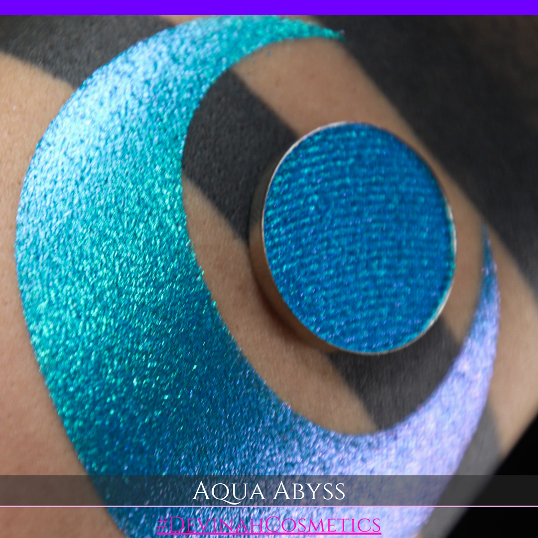 A round eyeshadow pan labeled 'AQUA ABYSS' made by Devinah Cosmetics, placed over a large crescent moon swatch of the same shimmering shifty eyeshadow on skin. The eyeshadow has a duochrome multichrome shift between colors lime green, blue, teal, pink.