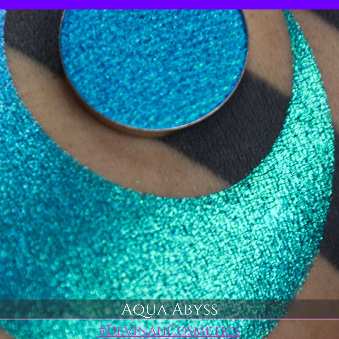 A round eyeshadow pan labeled 'AQUA ABYSS' made by Devinah Cosmetics, placed over a large crescent moon swatch of the same shimmering shifty eyeshadow on skin. The eyeshadow has a duochrome multichrome shift between colors lime green, blue, teal, pink.