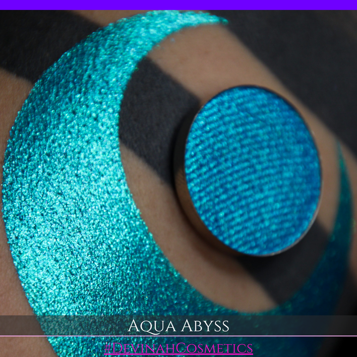 A round eyeshadow pan labeled 'AQUA ABYSS' made by Devinah Cosmetics, placed over a large crescent moon swatch of the same shimmering shifty eyeshadow on skin. The eyeshadow has a duochrome multichrome shift between colors lime green, blue, teal, pink.