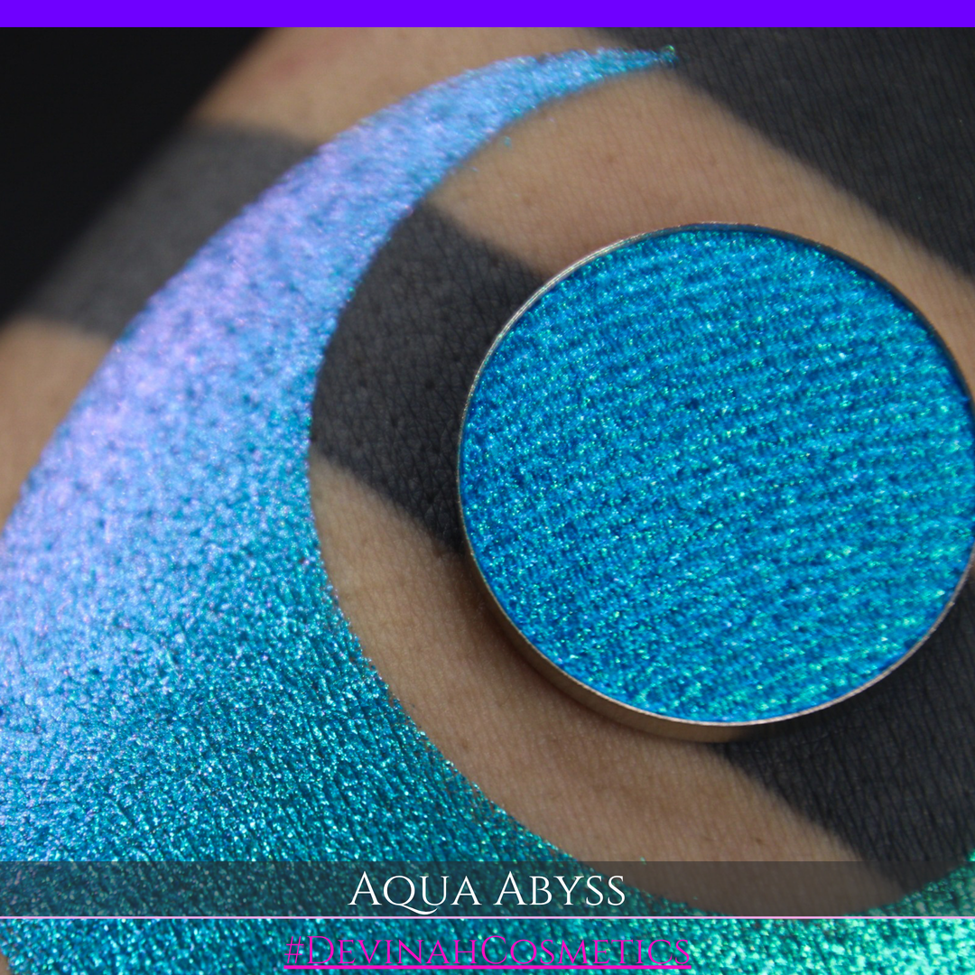 A round eyeshadow pan labeled 'AQUA ABYSS' made by Devinah Cosmetics, placed over a large crescent moon swatch of the same shimmering shifty eyeshadow on skin. The eyeshadow has a duochrome multichrome shift between colors lime green, blue, teal, pink.