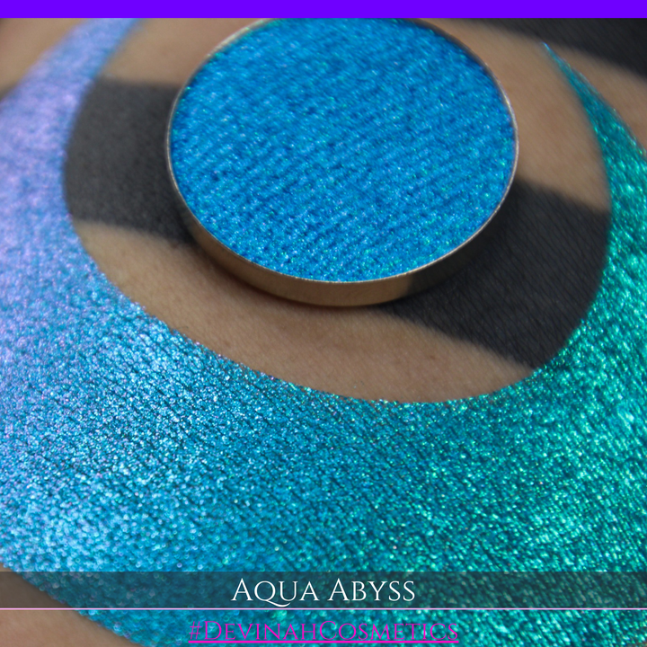 A round eyeshadow pan labeled 'AQUA ABYSS' made by Devinah Cosmetics, placed over a large crescent moon swatch of the same shimmering shifty eyeshadow on skin. The eyeshadow has a duochrome multichrome shift between colors lime green, blue, teal, pink.