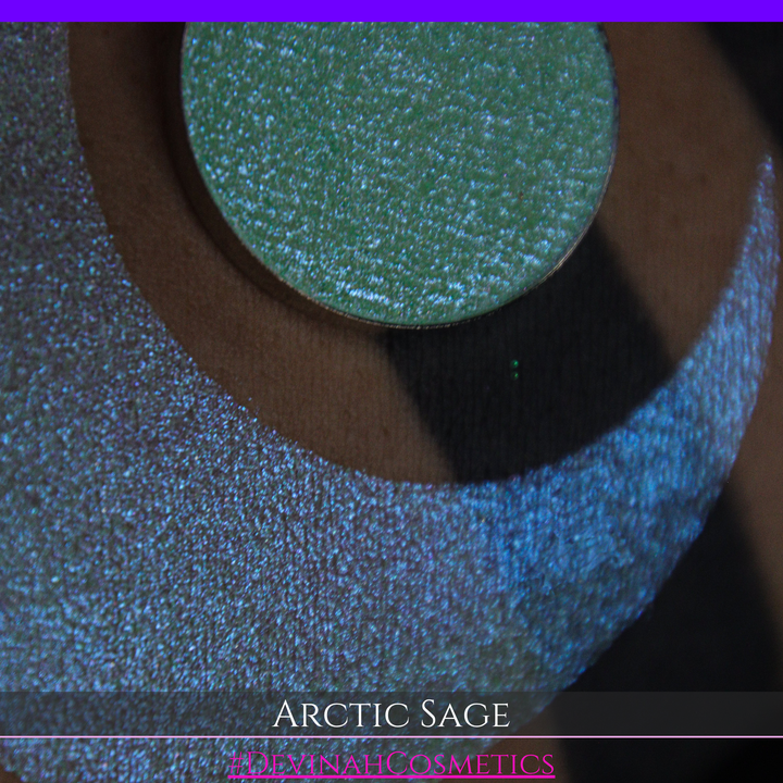 A close-up of a person's arm showing a swatch of shimmering eyeshadow and a circular eyeshadow pan labeled 'Arctic Sage' by Devinah Cosmetics, placed over a large crescent-shaped makeup swatch on skin.