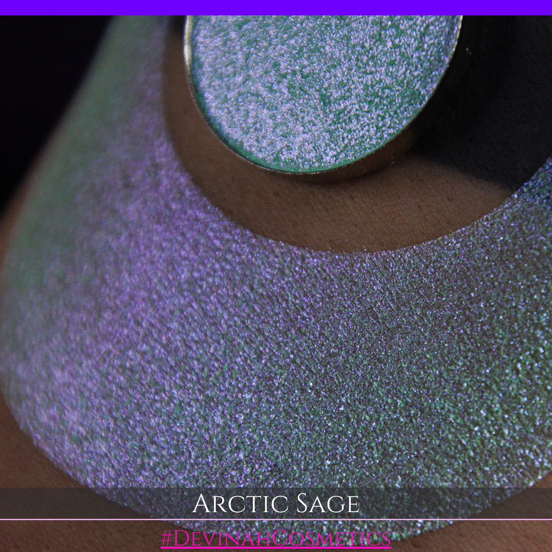 A close-up of a person's arm showing a swatch of shimmering eyeshadow and a circular eyeshadow pan labeled 'Arctic Sage' by Devinah Cosmetics, placed over a large crescent-shaped makeup swatch on skin.