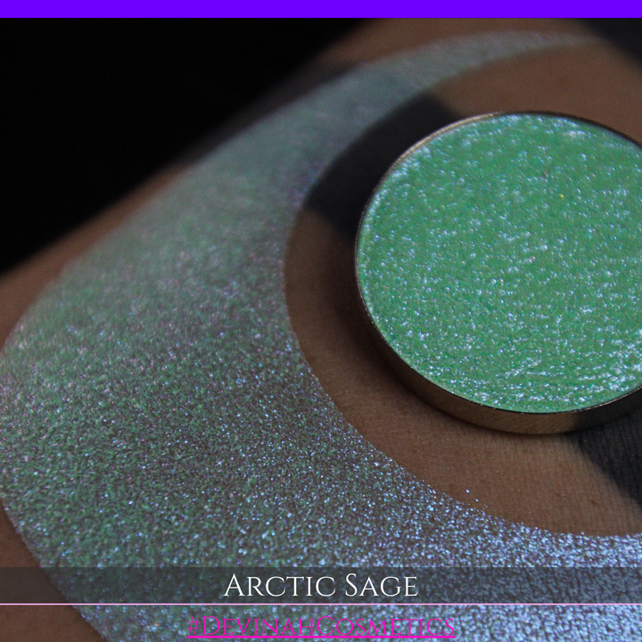 A close-up of a person's arm showing a swatch of shimmering eyeshadow and a circular eyeshadow pan labeled 'Arctic Sage' by Devinah Cosmetics, placed over a large crescent-shaped makeup swatch on skin.