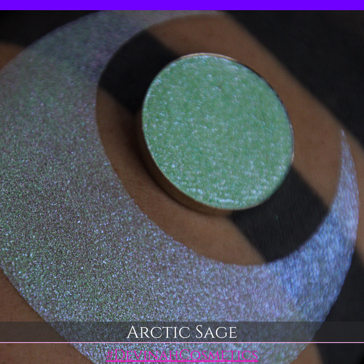 A close-up of a person's arm showing a swatch of shimmering eyeshadow and a circular eyeshadow pan labeled 'Arctic Sage' by Devinah Cosmetics, placed over a large crescent-shaped makeup swatch on skin.