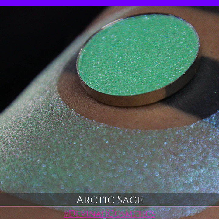 A close-up of a person's arm showing a swatch of shimmering eyeshadow and a circular eyeshadow pan labeled 'Arctic Sage' by Devinah Cosmetics, placed over a large crescent-shaped makeup swatch on skin.