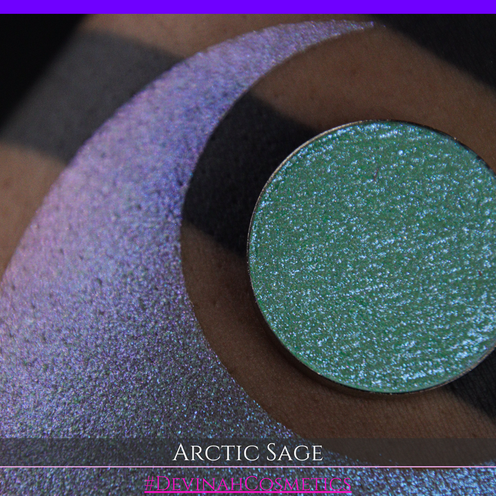A close-up of a person's arm showing a swatch of shimmering eyeshadow and a circular eyeshadow pan labeled 'Arctic Sage' by Devinah Cosmetics, placed over a large crescent-shaped makeup swatch on skin.