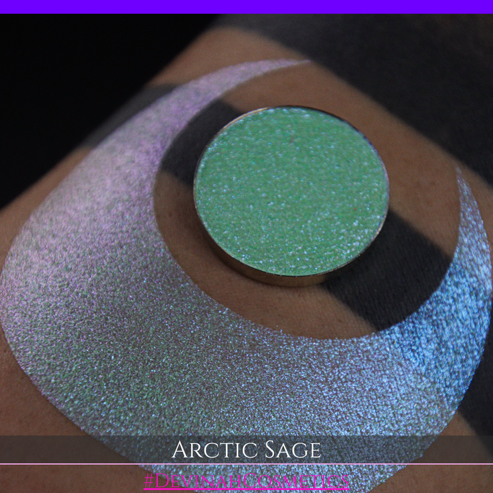 A close-up of a person's arm showing a swatch of shimmering eyeshadow and a circular eyeshadow pan labeled 'Arctic Sage' by Devinah Cosmetics, placed over a large crescent-shaped makeup swatch on skin.