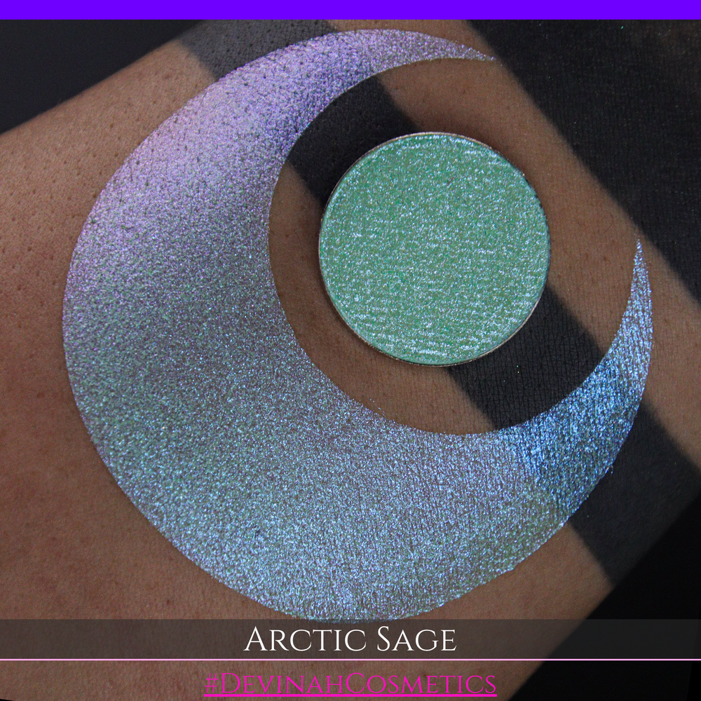 A close-up of a person's arm showing a swatch of shimmering eyeshadow and a circular eyeshadow pan labeled 'Arctic Sage' by Devinah Cosmetics, placed over a large crescent-shaped makeup swatch on skin.