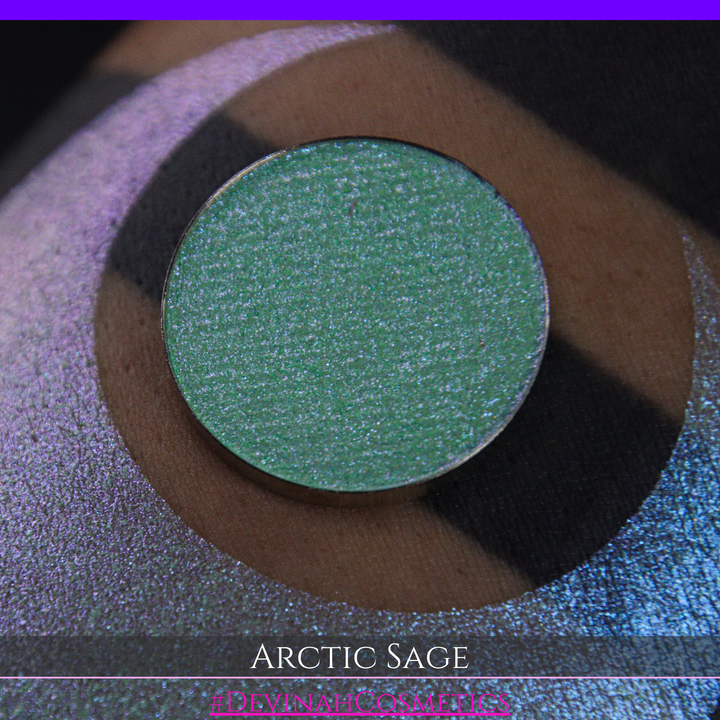 A close-up of a person's arm showing a swatch of shimmering eyeshadow and a circular eyeshadow pan labeled 'Arctic Sage' by Devinah Cosmetics, placed over a large crescent-shaped makeup swatch on skin.