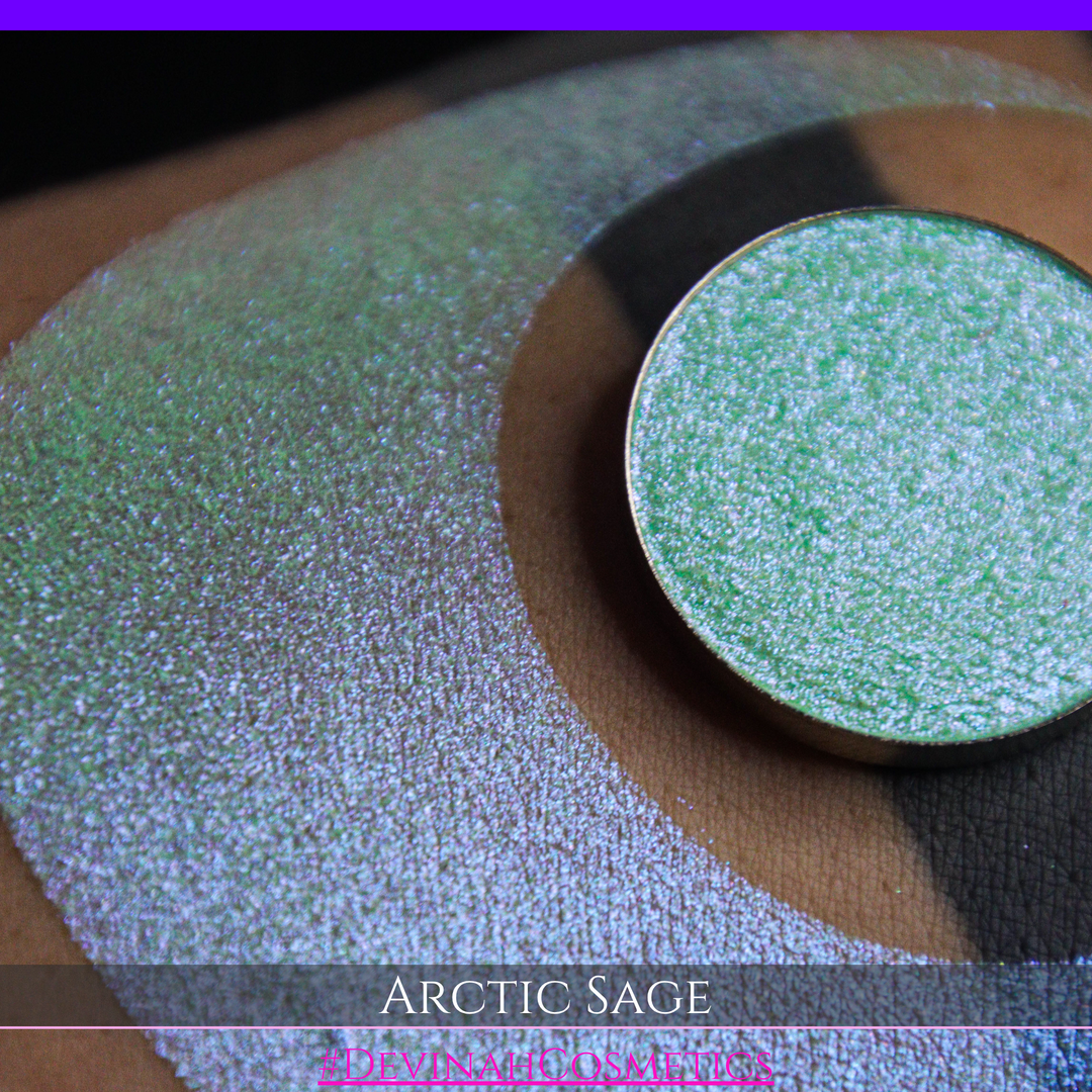 A close-up of a person's arm showing a swatch of shimmering eyeshadow and a circular eyeshadow pan labeled 'Arctic Sage' by Devinah Cosmetics, placed over a large crescent-shaped makeup swatch on skin.