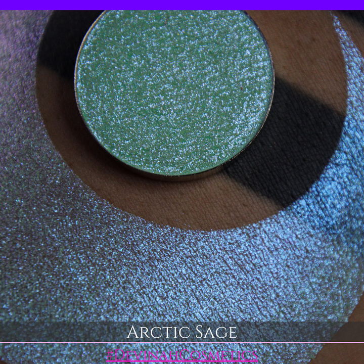 A close-up of a person's arm showing a swatch of shimmering eyeshadow and a circular eyeshadow pan labeled 'Arctic Sage' by Devinah Cosmetics, placed over a large crescent-shaped makeup swatch on skin.