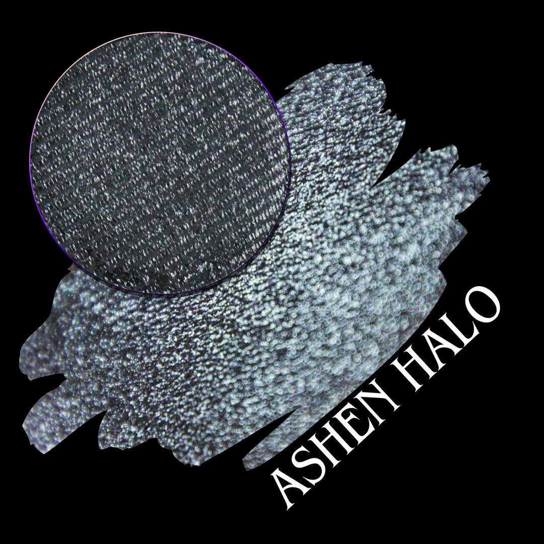 A round eyeshadow pan labeled 'ASHEN HALO' made by Devinah Cosmetics, placed over a large crescent moon swatch of the same shimmering shifty eyeshadow on skin. The eyeshadow has a duochrome multichrome shift between colors steel gray, grey, blue, green.