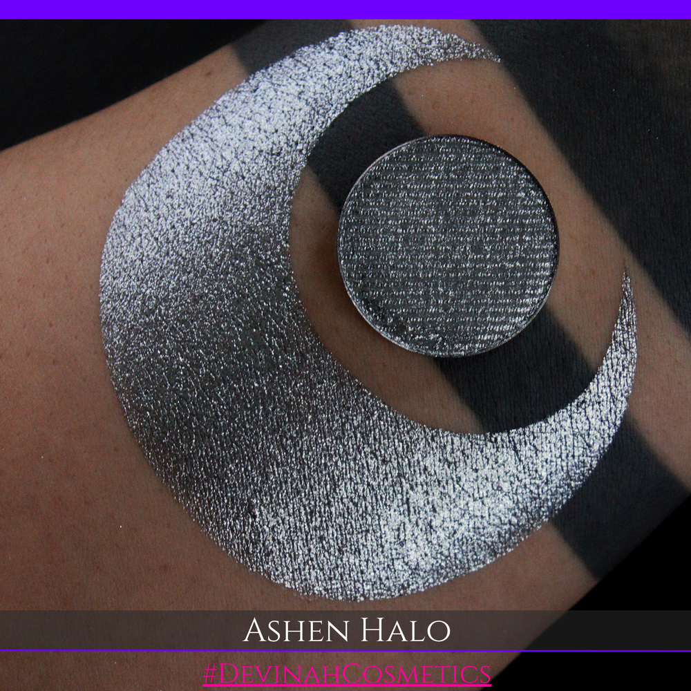 A round eyeshadow pan labeled 'ASHEN HALO' made by Devinah Cosmetics, placed over a large crescent moon swatch of the same shimmering shifty eyeshadow on skin. The eyeshadow has a duochrome multichrome shift between colors steel gray, grey, blue, green.