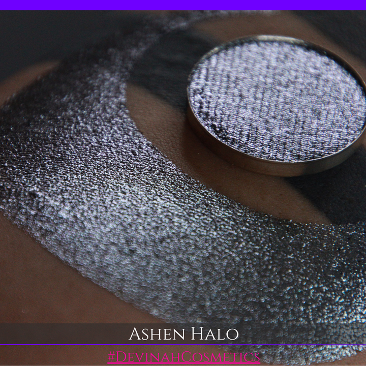 A round eyeshadow pan labeled 'ASHEN HALO' made by Devinah Cosmetics, placed over a large crescent moon swatch of the same shimmering shifty eyeshadow on skin. The eyeshadow has a duochrome multichrome shift between colors steel gray, grey, blue, green.