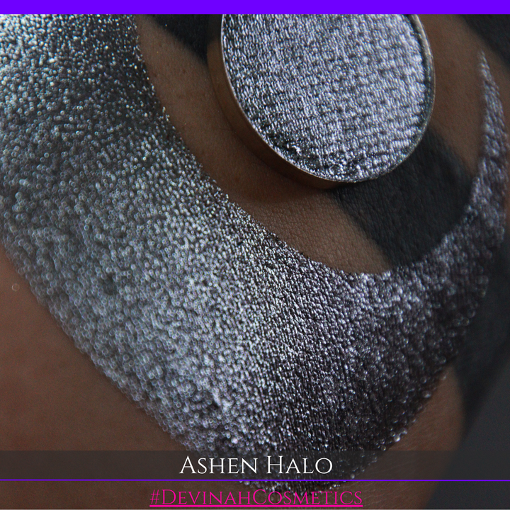 A round eyeshadow pan labeled 'ASHEN HALO' made by Devinah Cosmetics, placed over a large crescent moon swatch of the same shimmering shifty eyeshadow on skin. The eyeshadow has a duochrome multichrome shift between colors steel gray, grey, blue, green.