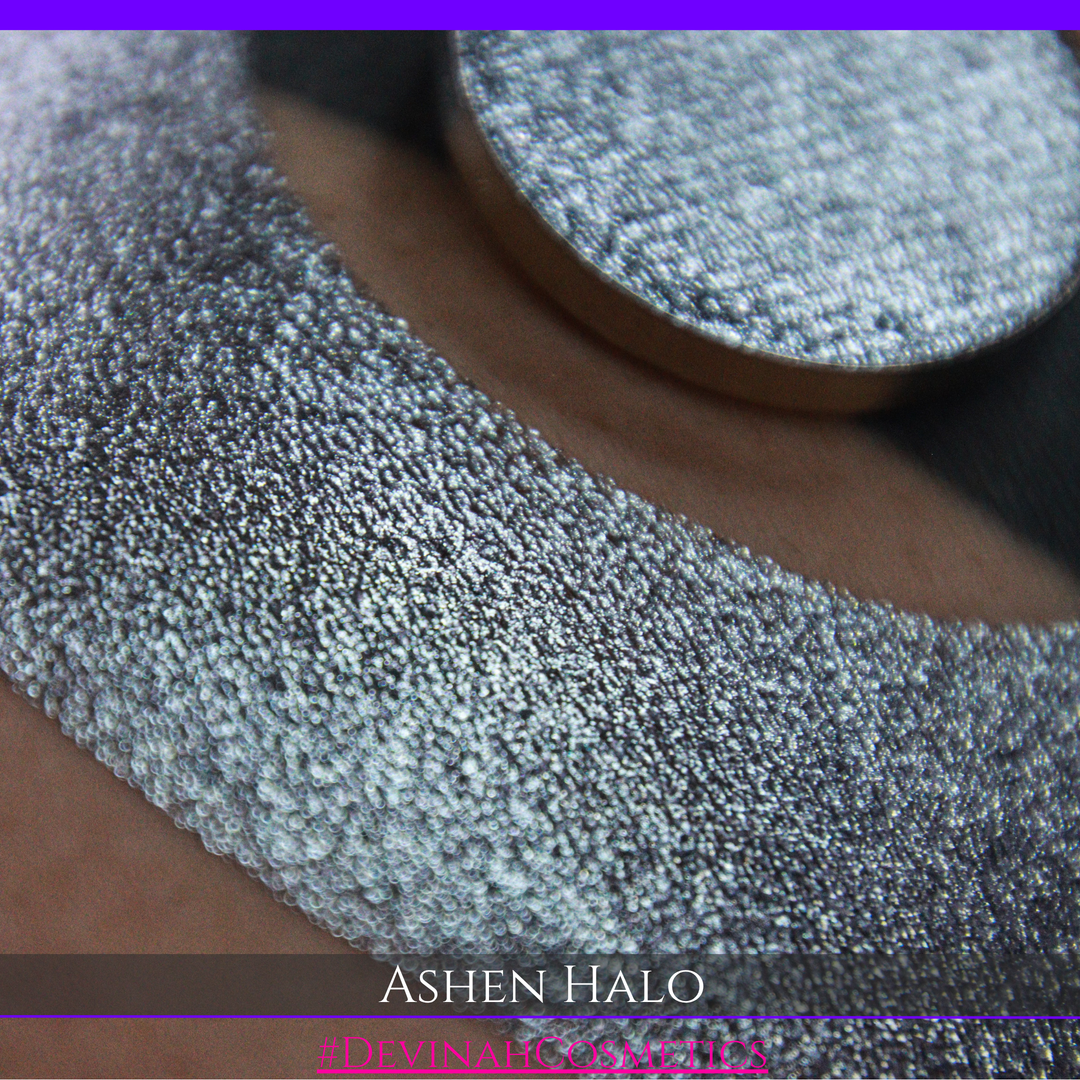 A round eyeshadow pan labeled 'ASHEN HALO' made by Devinah Cosmetics, placed over a large crescent moon swatch of the same shimmering shifty eyeshadow on skin. The eyeshadow has a duochrome multichrome shift between colors steel gray, grey, blue, green.