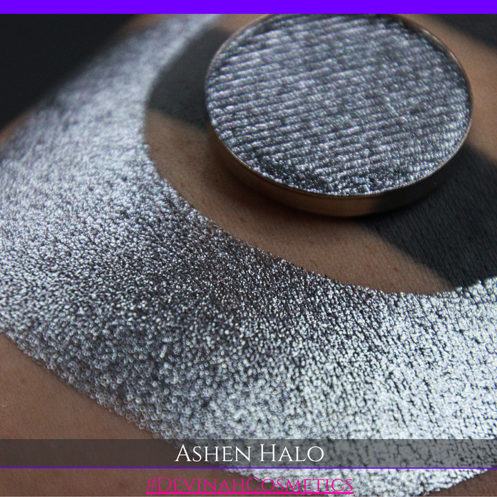 A round eyeshadow pan labeled 'ASHEN HALO' made by Devinah Cosmetics, placed over a large crescent moon swatch of the same shimmering shifty eyeshadow on skin. The eyeshadow has a duochrome multichrome shift between colors steel gray, grey, blue, green.