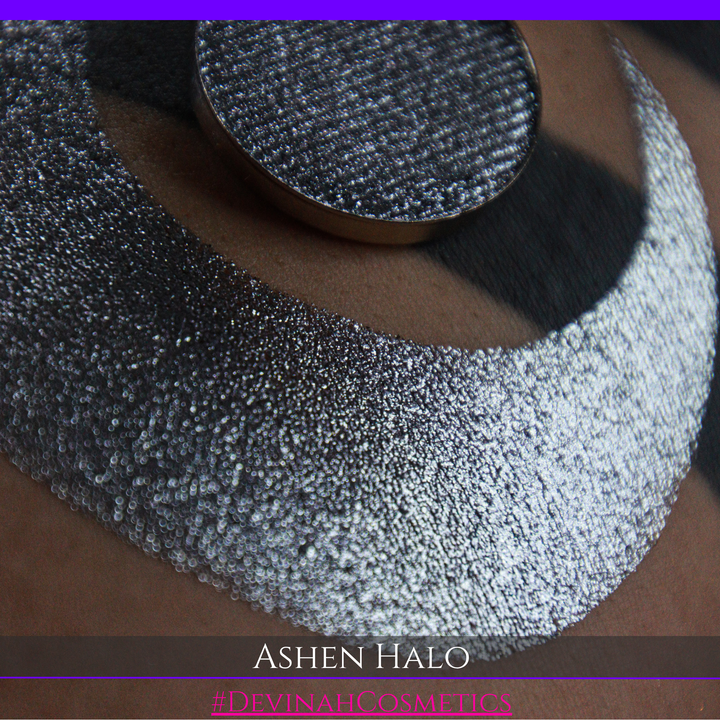 A round eyeshadow pan labeled 'ASHEN HALO' made by Devinah Cosmetics, placed over a large crescent moon swatch of the same shimmering shifty eyeshadow on skin. The eyeshadow has a duochrome multichrome shift between colors steel gray, grey, blue, green.