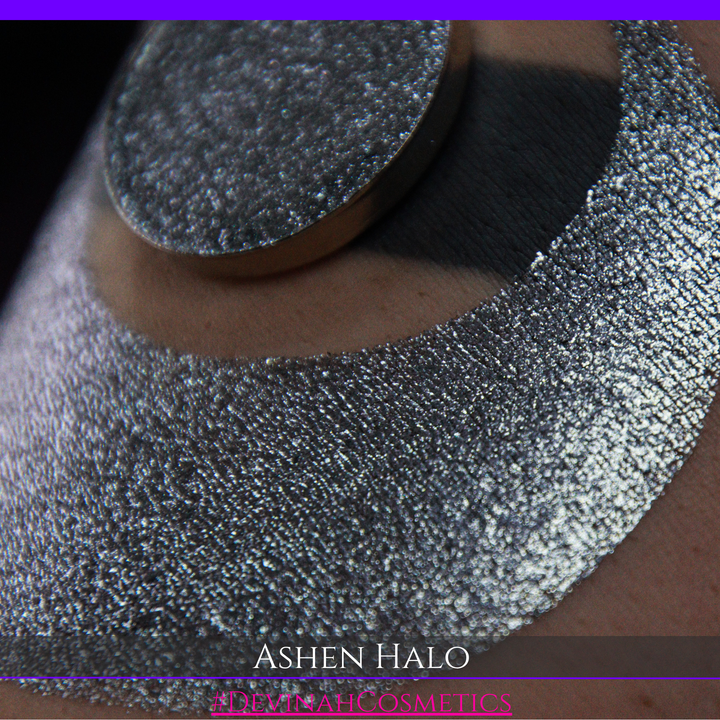 A round eyeshadow pan labeled 'ASHEN HALO' made by Devinah Cosmetics, placed over a large crescent moon swatch of the same shimmering shifty eyeshadow on skin. The eyeshadow has a duochrome multichrome shift between colors steel gray, grey, blue, green.