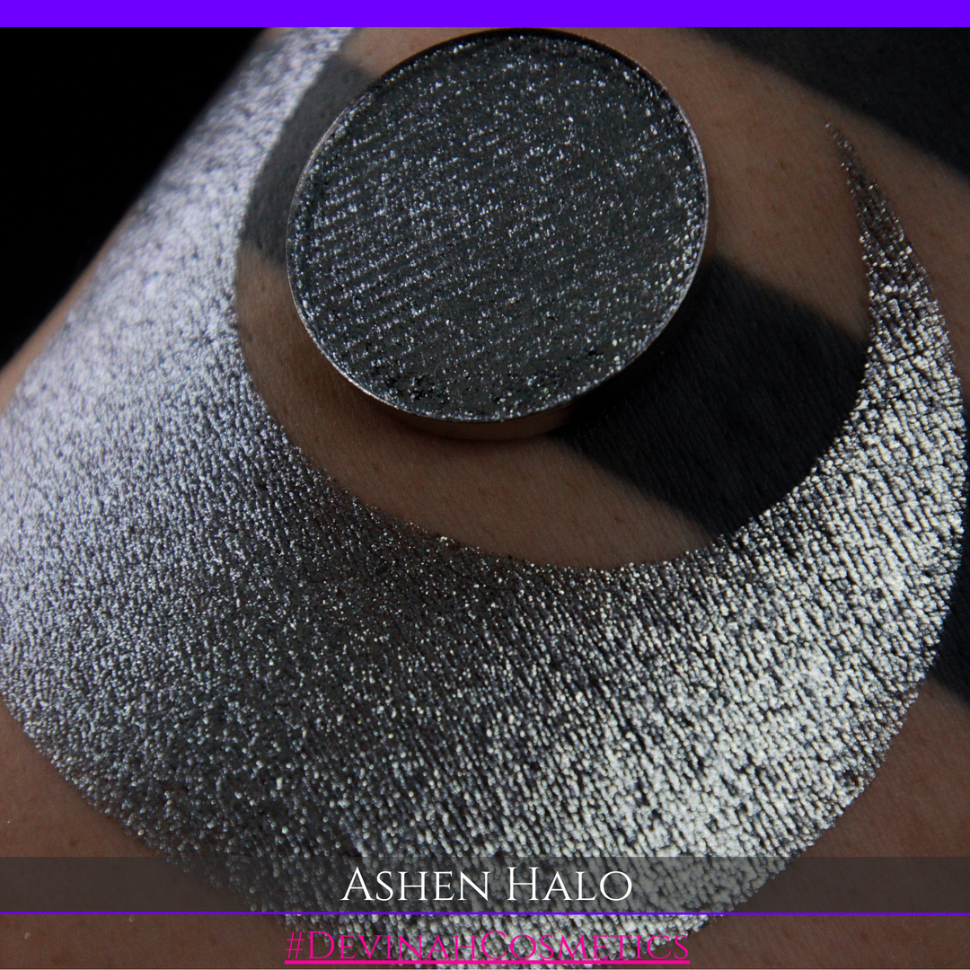 A round eyeshadow pan labeled 'ASHEN HALO' made by Devinah Cosmetics, placed over a large crescent moon swatch of the same shimmering shifty eyeshadow on skin. The eyeshadow has a duochrome multichrome shift between colors steel gray, grey, blue, green.