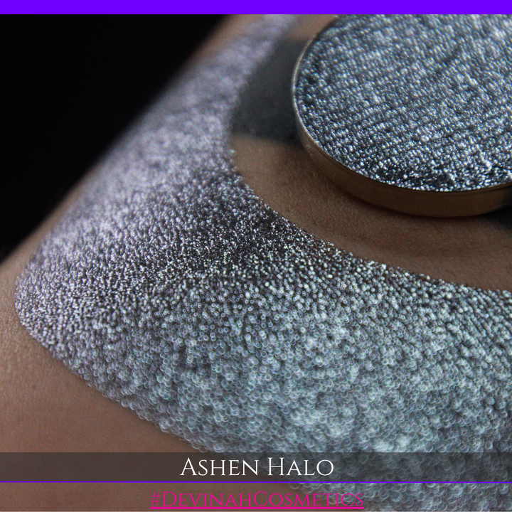 A round eyeshadow pan labeled 'ASHEN HALO' made by Devinah Cosmetics, placed over a large crescent moon swatch of the same shimmering shifty eyeshadow on skin. The eyeshadow has a duochrome multichrome shift between colors steel gray, grey, blue, green.