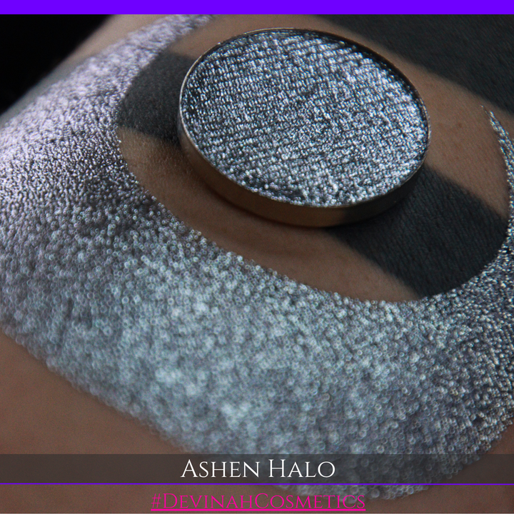 A round eyeshadow pan labeled 'ASHEN HALO' made by Devinah Cosmetics, placed over a large crescent moon swatch of the same shimmering shifty eyeshadow on skin. The eyeshadow has a duochrome multichrome shift between colors steel gray, grey, blue, green.