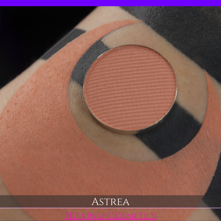 A close-up of a person's arm with a large peach-toned makeup swatch and a matching round eyeshadow makeup pan labeled ASTREA, with the text #DEVINAHCOSMETICS at the bottom.