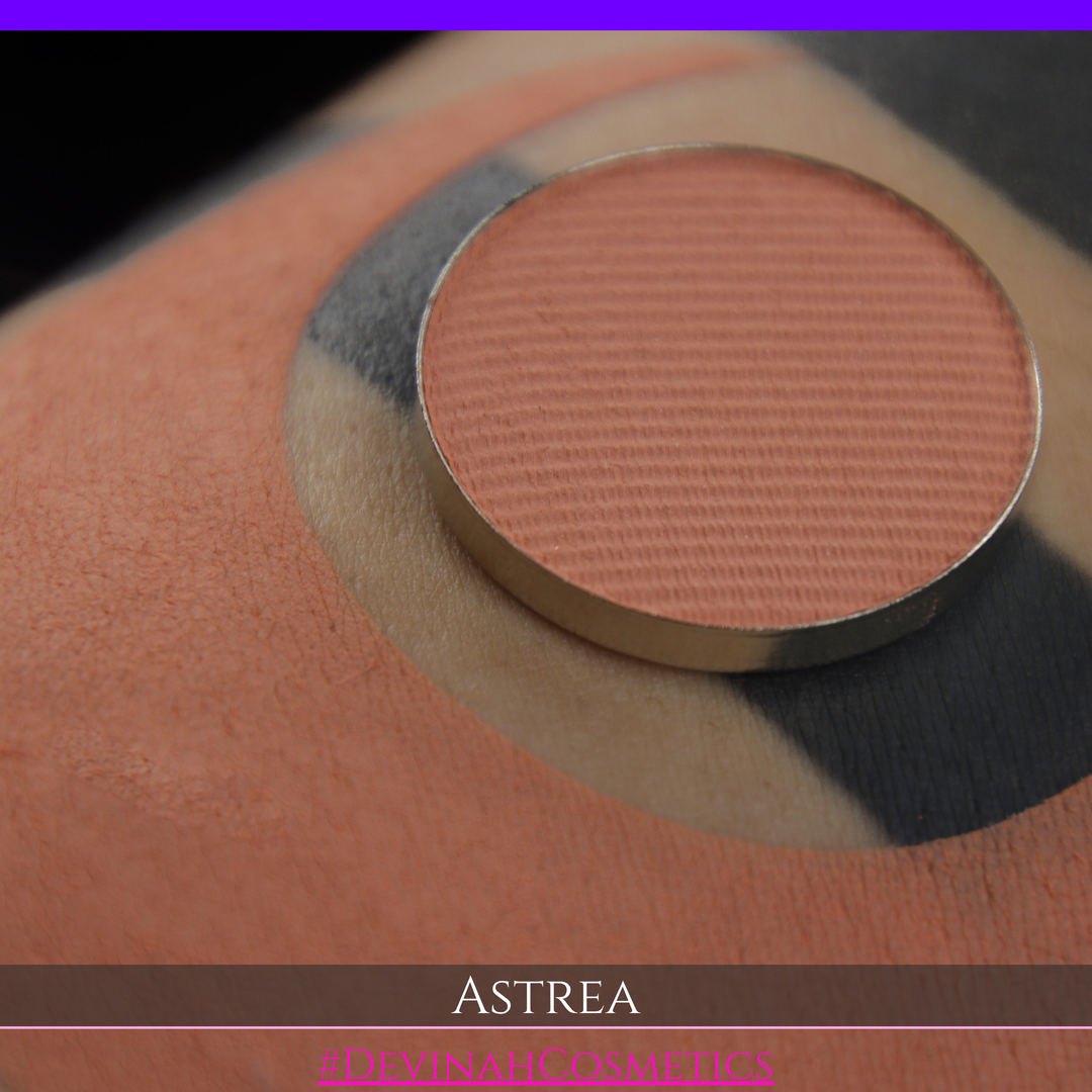 A close-up of a person's arm with a large peach-toned makeup swatch and a matching round eyeshadow makeup pan labeled ASTREA, with the text #DEVINAHCOSMETICS at the bottom.